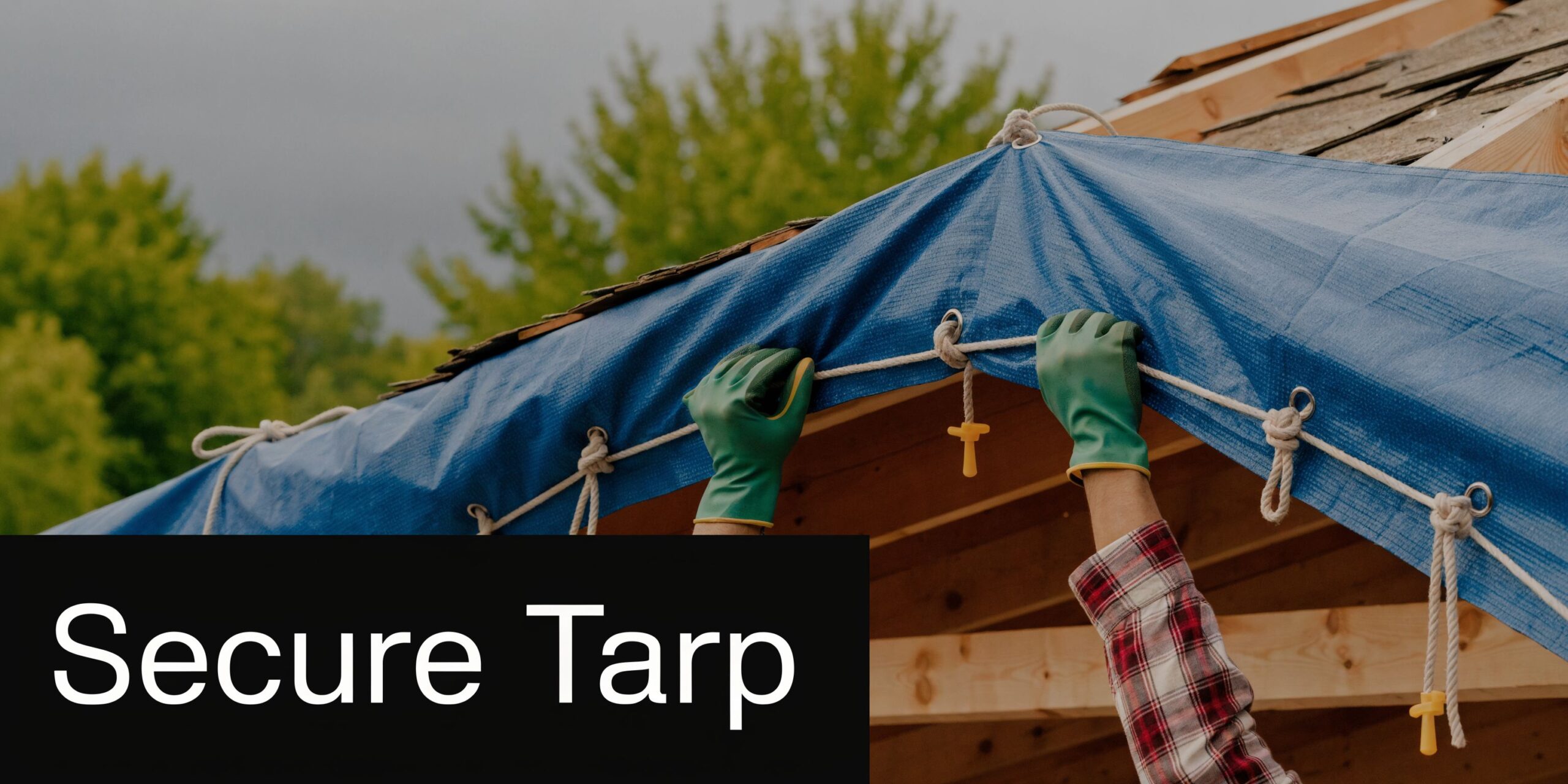A person in work gloves secures a blue tarp over a wooden roof frame during construction.