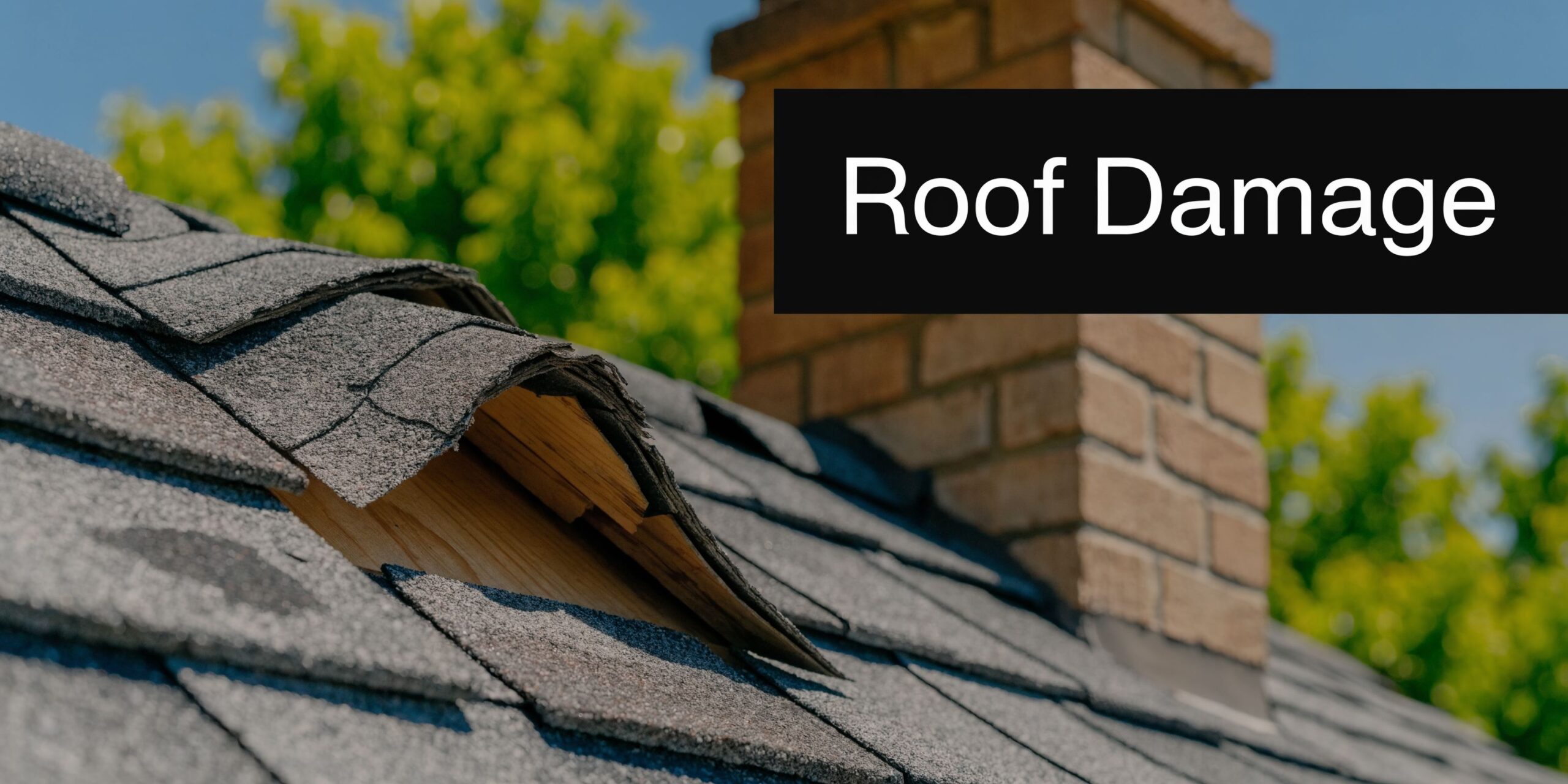 A close-up view of damaged asphalt roof shingles with exposed wood decking near a brick chimney.