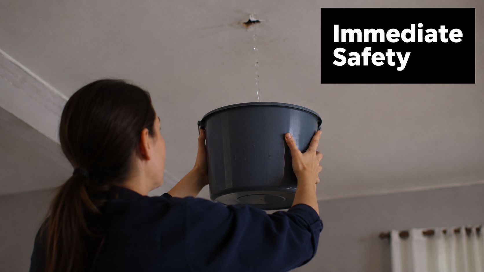 A woman holding a grey bucket under a leaking ceiling to catch dripping water from a hole.
